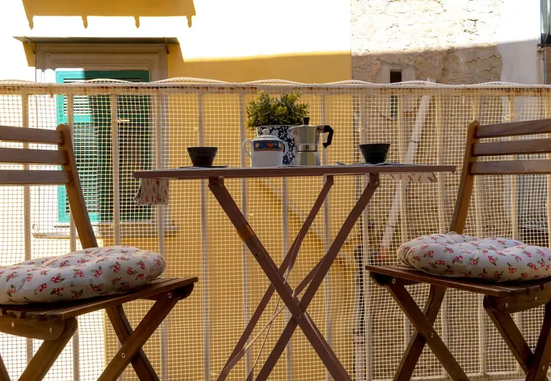 Il balcone: due sedie di legno con i cuscini a fiori, il tavolino con la moka e le tazze, di fronte la facciata gialla con le persiane verdi.